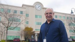 Rahim Opeyany, an older bald man with glasses and a blue button up short, stands in front of a large cream colored building with many windows and a large clock.