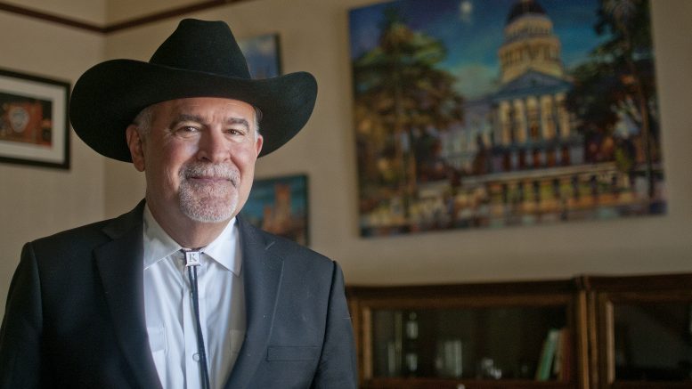 Evan Edgar, a white haired man in a black cowboy hat and bolo tie, stands in his office and smiles for the camera in front of a painting of the Capitol.