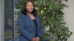 Janay Eustace, a black woman in a navy blazer, stands outside and smiles for the camera