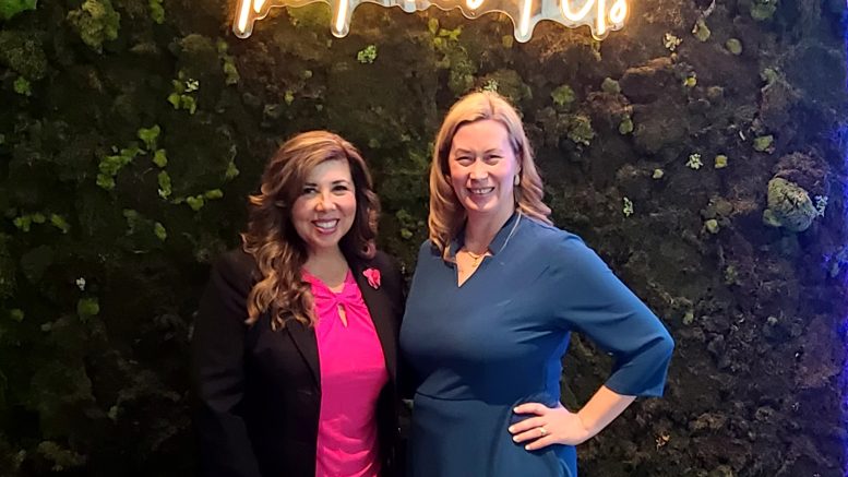 Two women standing next to each other smiling with a neon sign in the background that says, "the power of us".