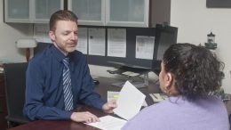 Man dressed in business attire sitting across from a woman at a desk talking.