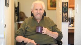 Man sitting in a motorized wheel chair in his house, smiling, with a coffee mug in his hand.