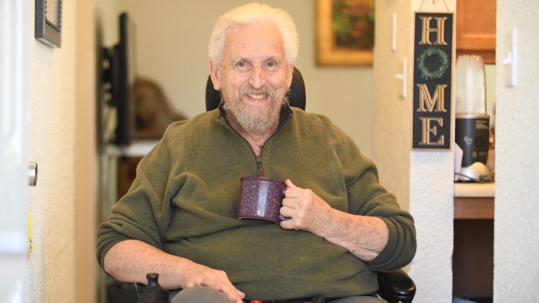 Man sitting in a motorized wheel chair in his house, smiling, with a coffee mug in his hand.