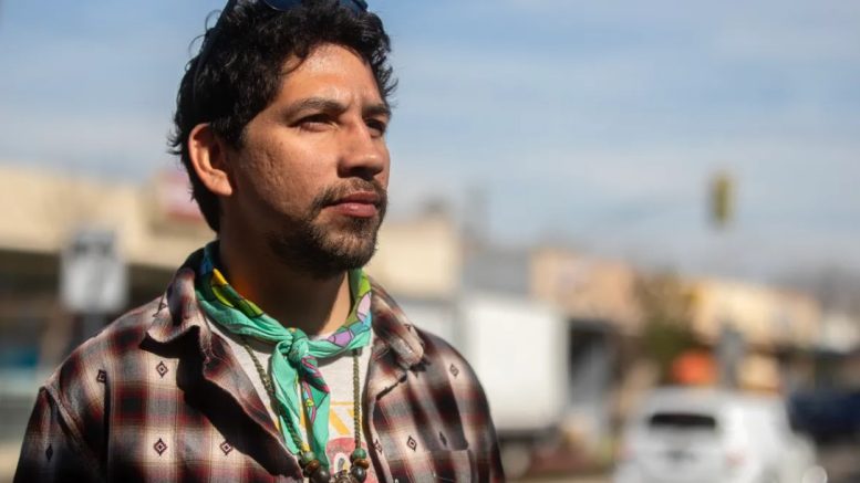 A person with short dark hair and facial hair stands outdoors, looking off into the distance with a focused expression. They wear a plaid shirt, a green bandana around their neck, and a beaded necklace, with sunglasses resting on their head. The background shows a softly blurred street scene with vehicles and buildings.