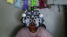A child, wearing a pink Strawberry shortcake shirt, sits on a chair with their eyes positioned in front of a metal Phoropter during an eye exam.