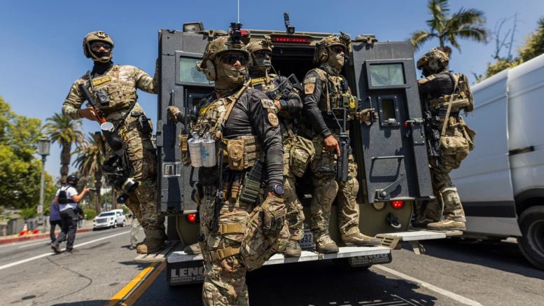 A group of heavily armed federal agents in full tactical gear emerge from the back of an armored vehicle labeled “LENCO ARMORED VEHICLES” on a sunlit urban street. Most wear camouflage fatigues, body armor, helmets, and goggles, with visible patches reading “POLICE” and insignias from U.S. Border Patrol and Homeland Security. Some agents carry rifles and one has zip cuffs and canisters clipped to their vest. A media photographer in a press vest films nearby. Palm trees and a clear blue sky suggest a Southern California location.