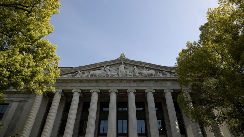 View looking up at a neoclassical building with tall columns and sculpted figures, framed by leafy trees against a clear blue sky.