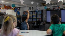 The back of elementary-aged students as they look towards a teacher standing at the front of a classroom. The photo focuses on a student wearing a blue shirt and raising their right hand in the air.