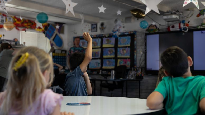 The back of elementary-aged students as they look towards a teacher standing at the front of a classroom. The photo focuses on a student wearing a blue shirt and raising their right hand in the air.